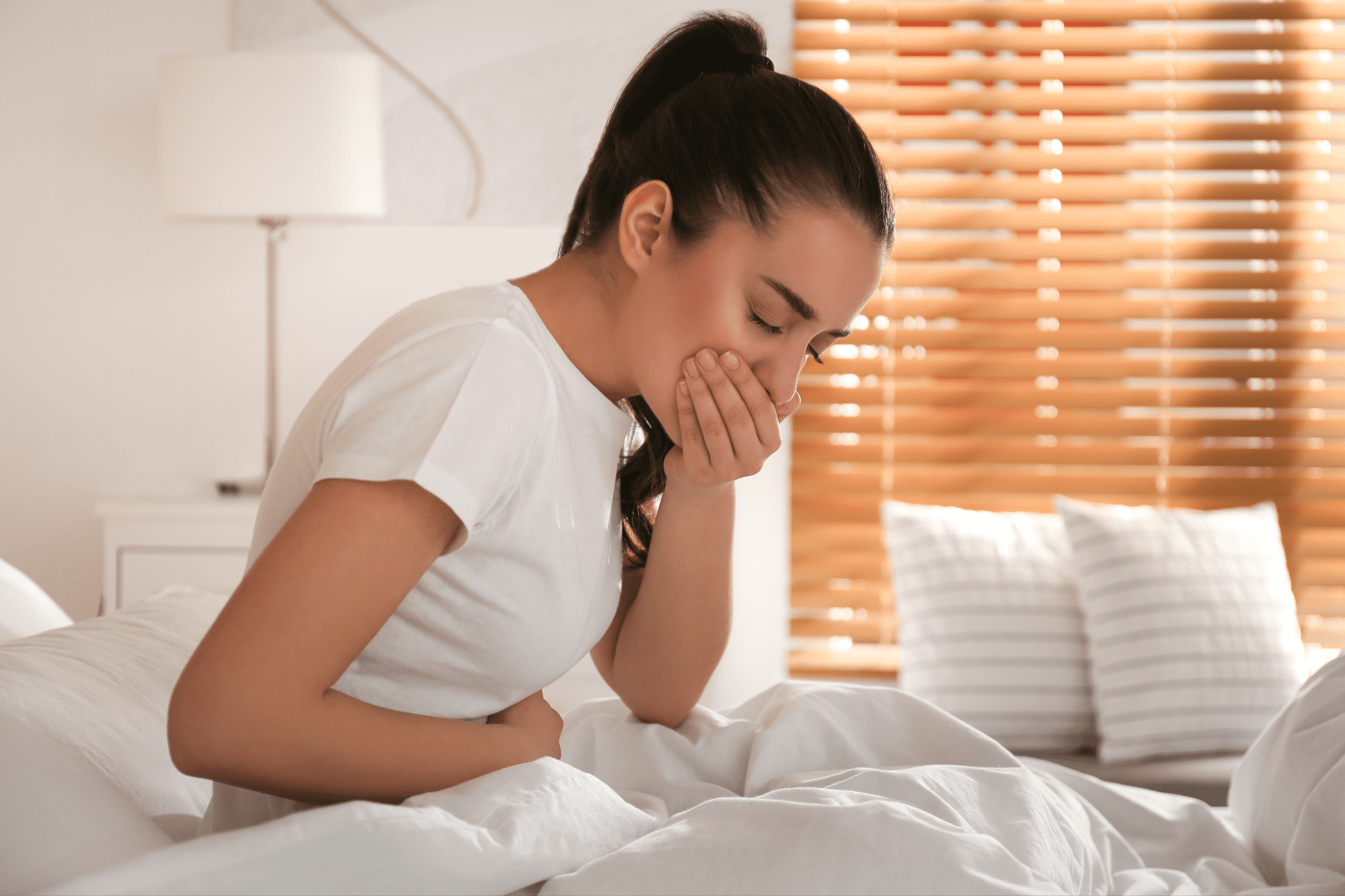 Woman sitting in bed with a hand clasped over her mouth and the other wrapped around her stomach